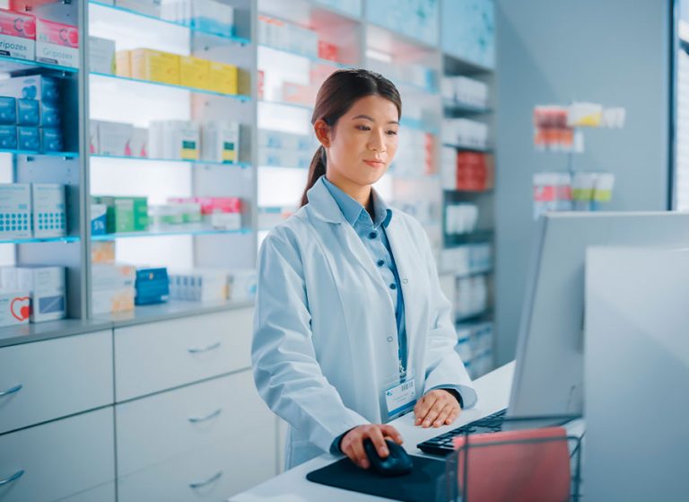 Mulher farmacêutica, mexendo em computador, dentro de uma farmácia, com medicamentos em prateleira