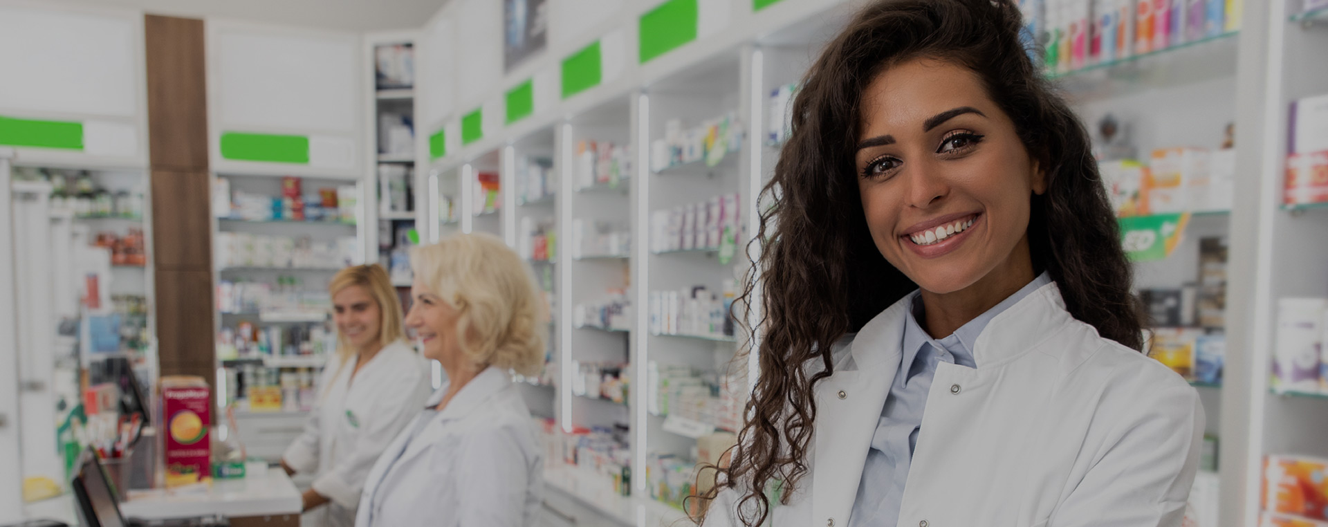 Mulher farmacêutica sorrindo em pé dentro de farmácia e duas outras atendentes atrás
